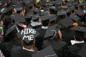 Crowd of grad students with caps on, one standing out with the words HIRE ME
